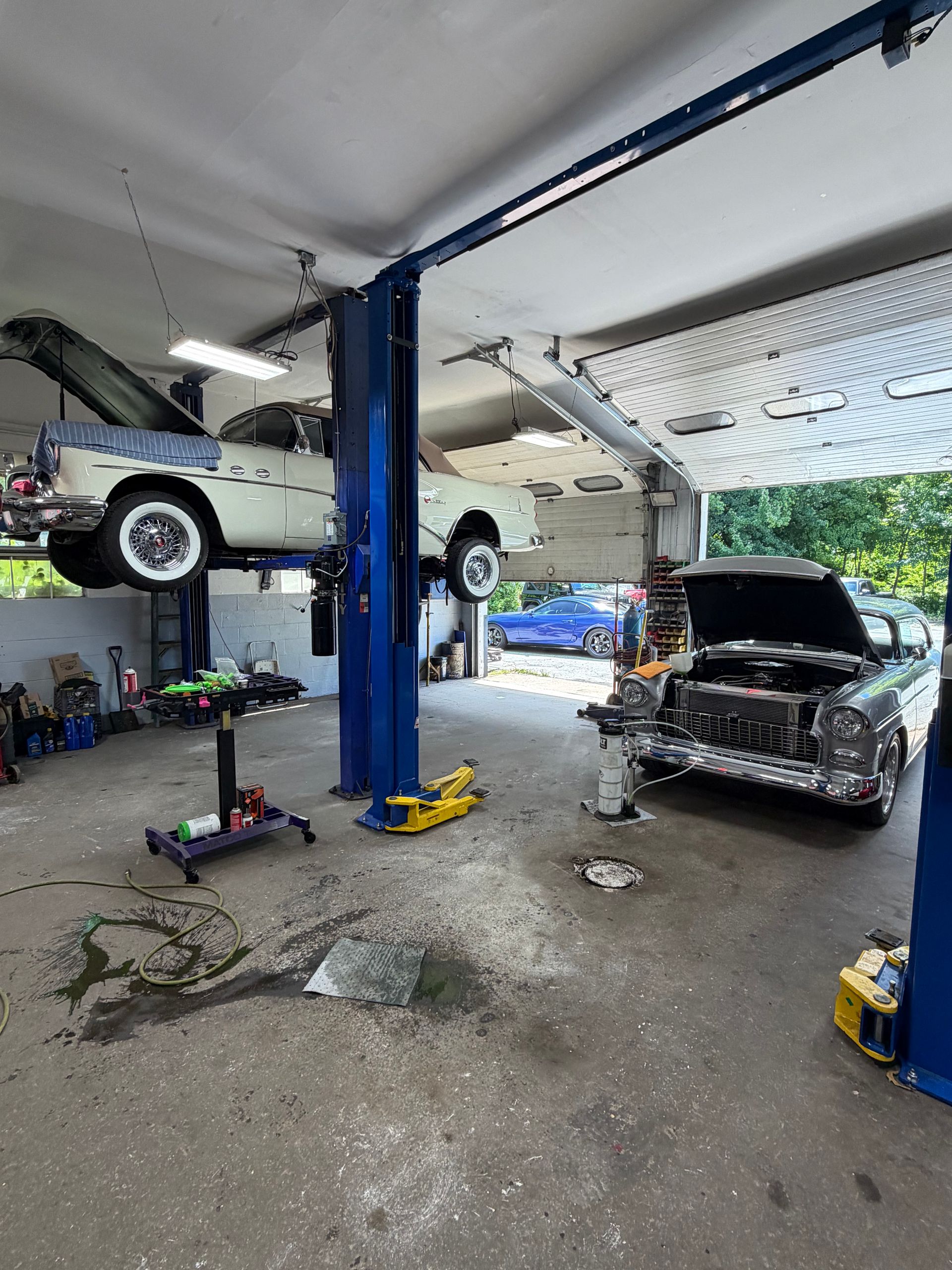 Car repair garage with two cars on lifts, one hood open, and tools scattered on the floor.