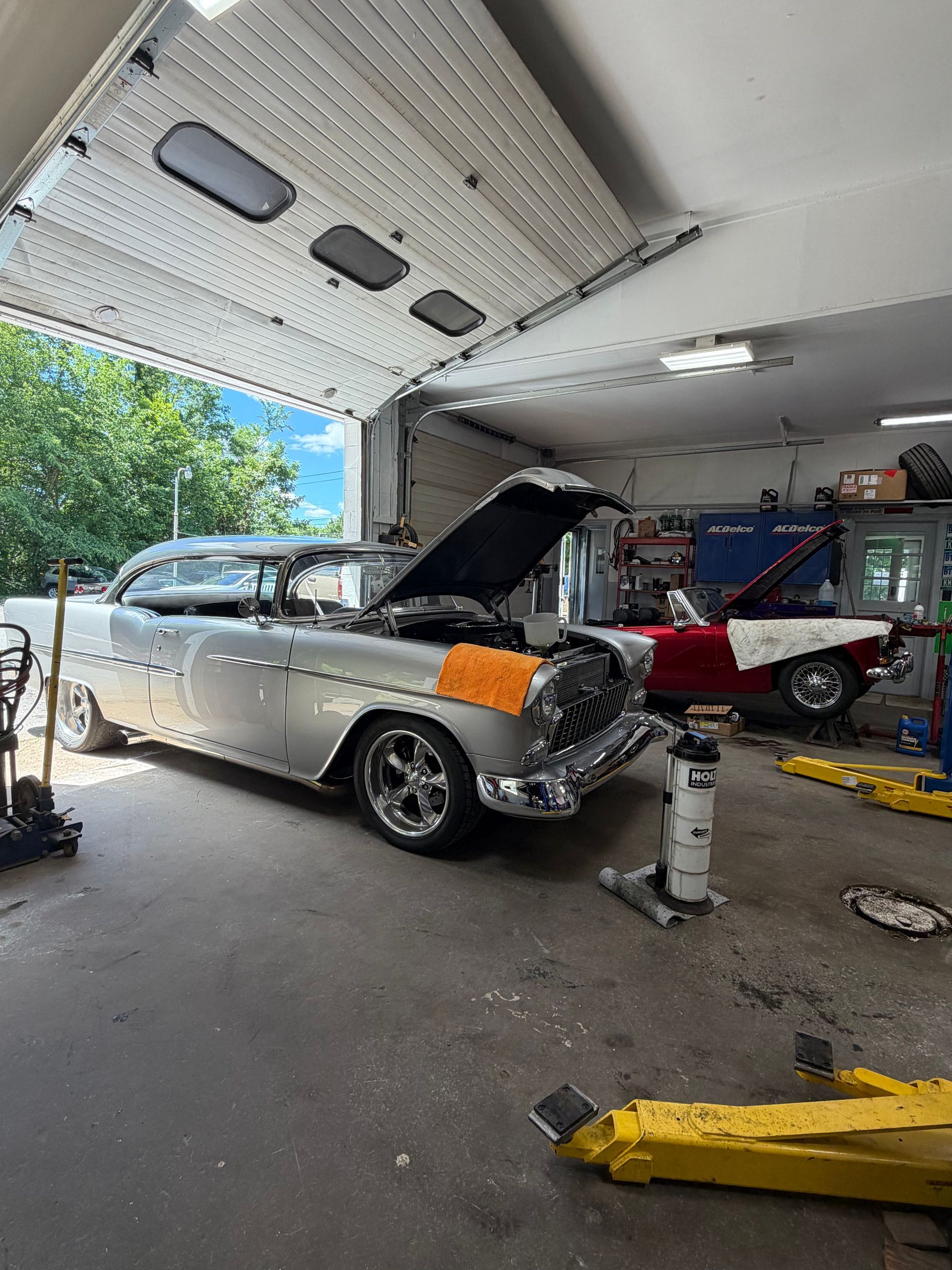 Silver car with hood open inside a garage, front bumper damaged and orange fender visible.