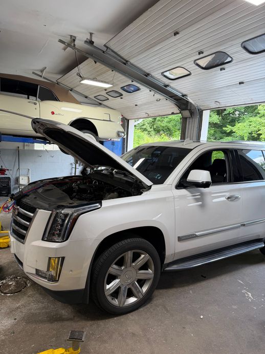 White SUV with hood open in a garage, parked beside a dark car under a lift.