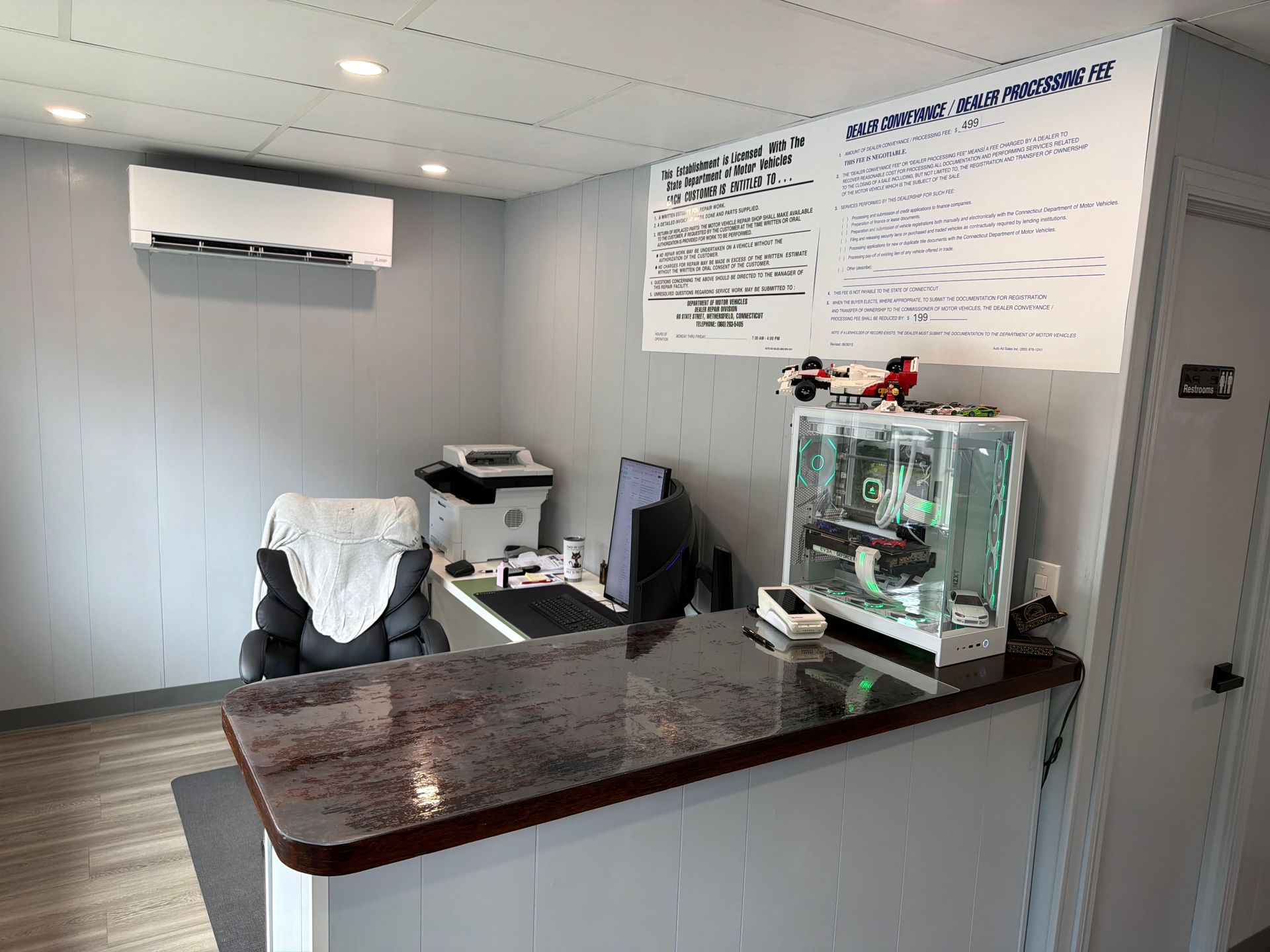 Small office reception area with a dark desk, wall AC, chair, printer, and glass display case.