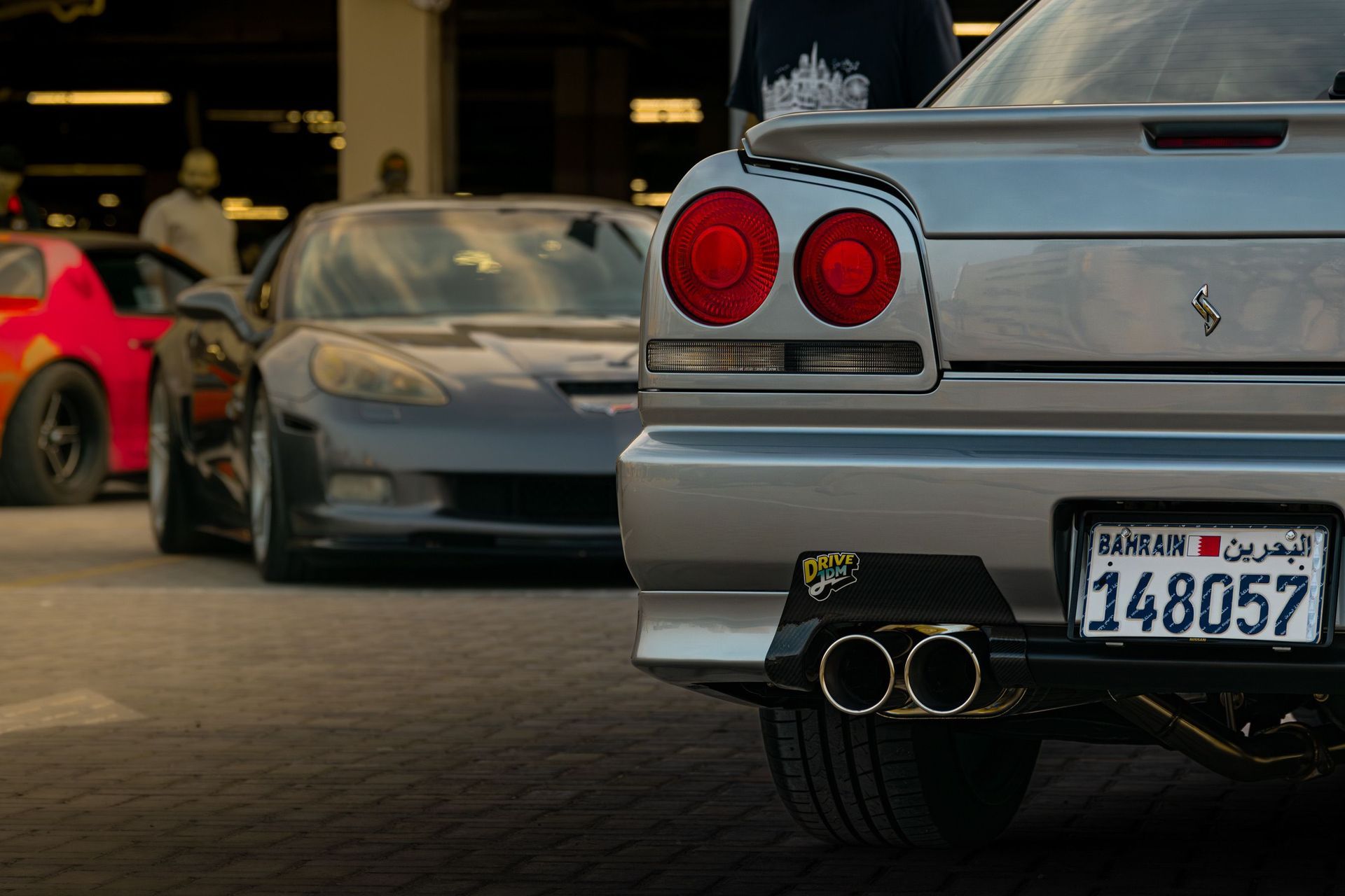 Rear view of a silver sports car with twin exhausts and a license plate, with other cars behind it in a garage