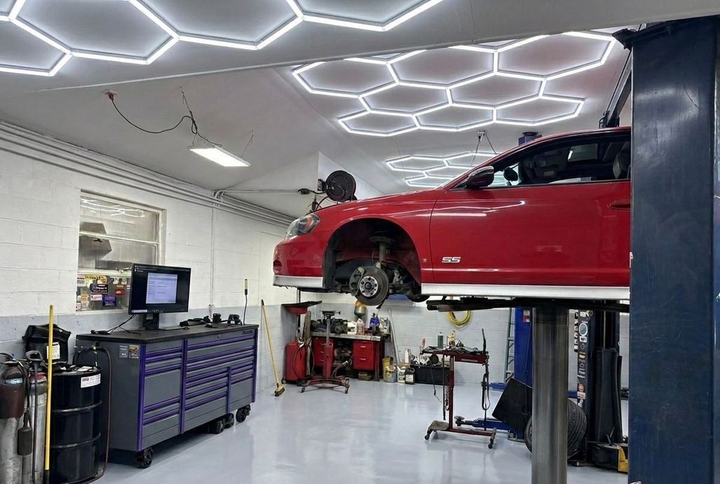 Red sports car on a lift in a bright auto repair garage with hexagon ceiling lights and tools below.