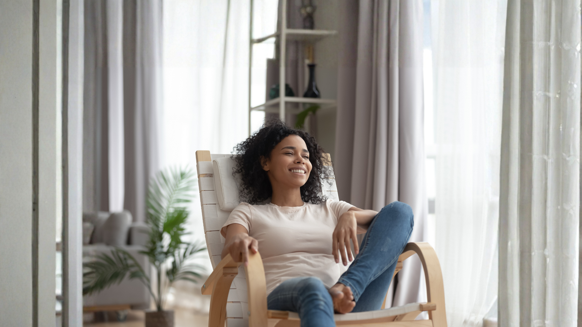 A smiling person relaxes in a wooden chair next to a large window with white curtains in a bright, modern living room.