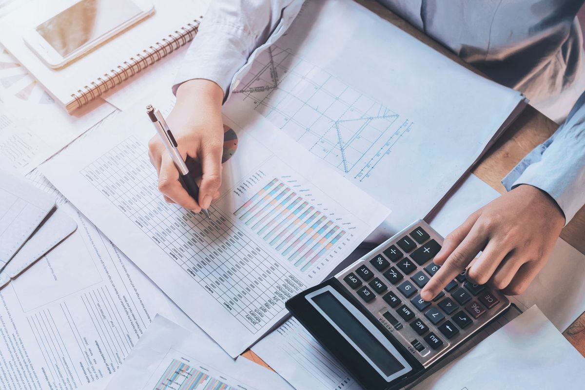 Hands using a calculator and writing on financial documents at a desk, sunlight.
