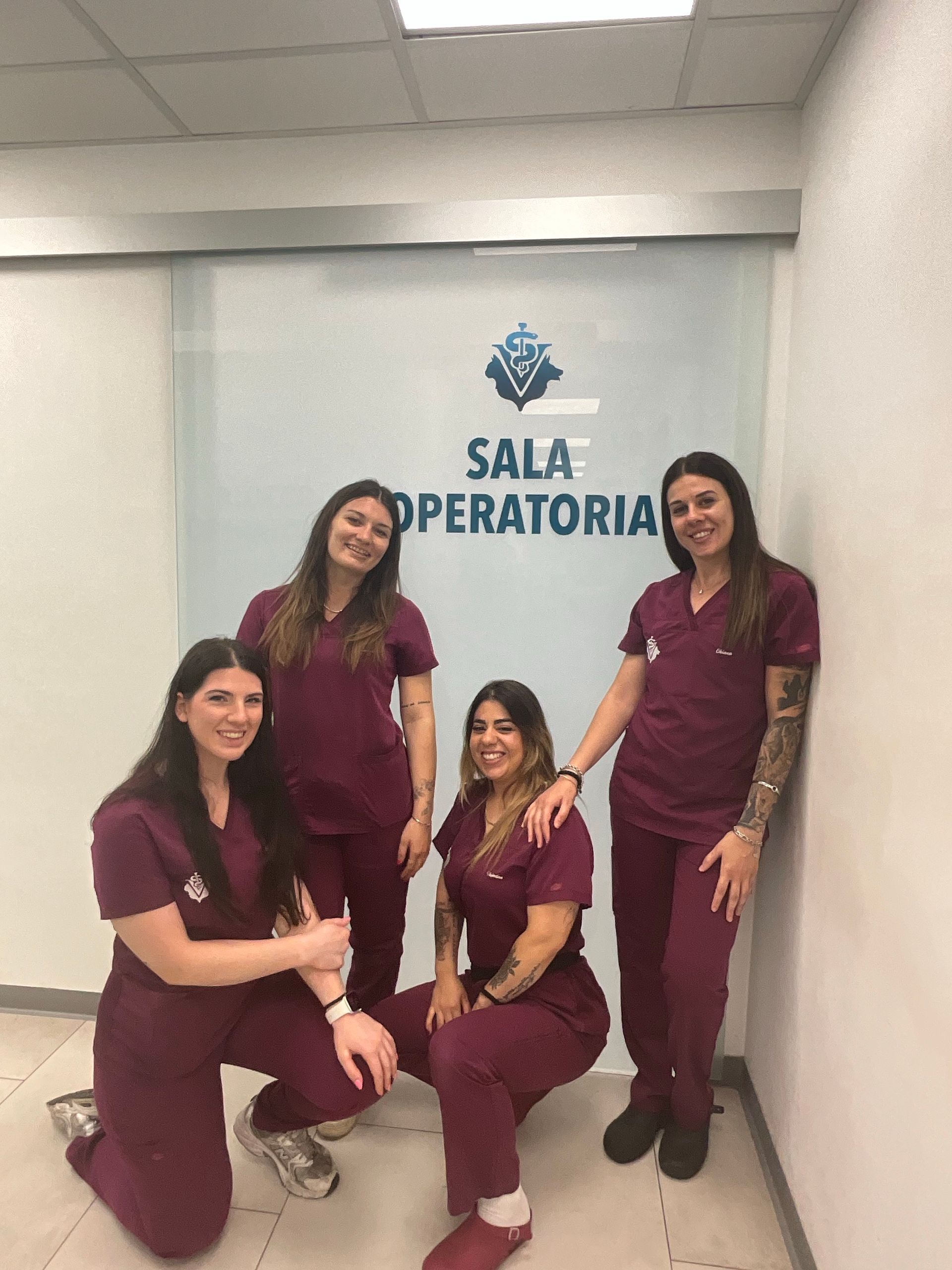 Quattro operatori sanitari in camice color bordeaux posano in un corridoio fuori da una stanza con la scritta 