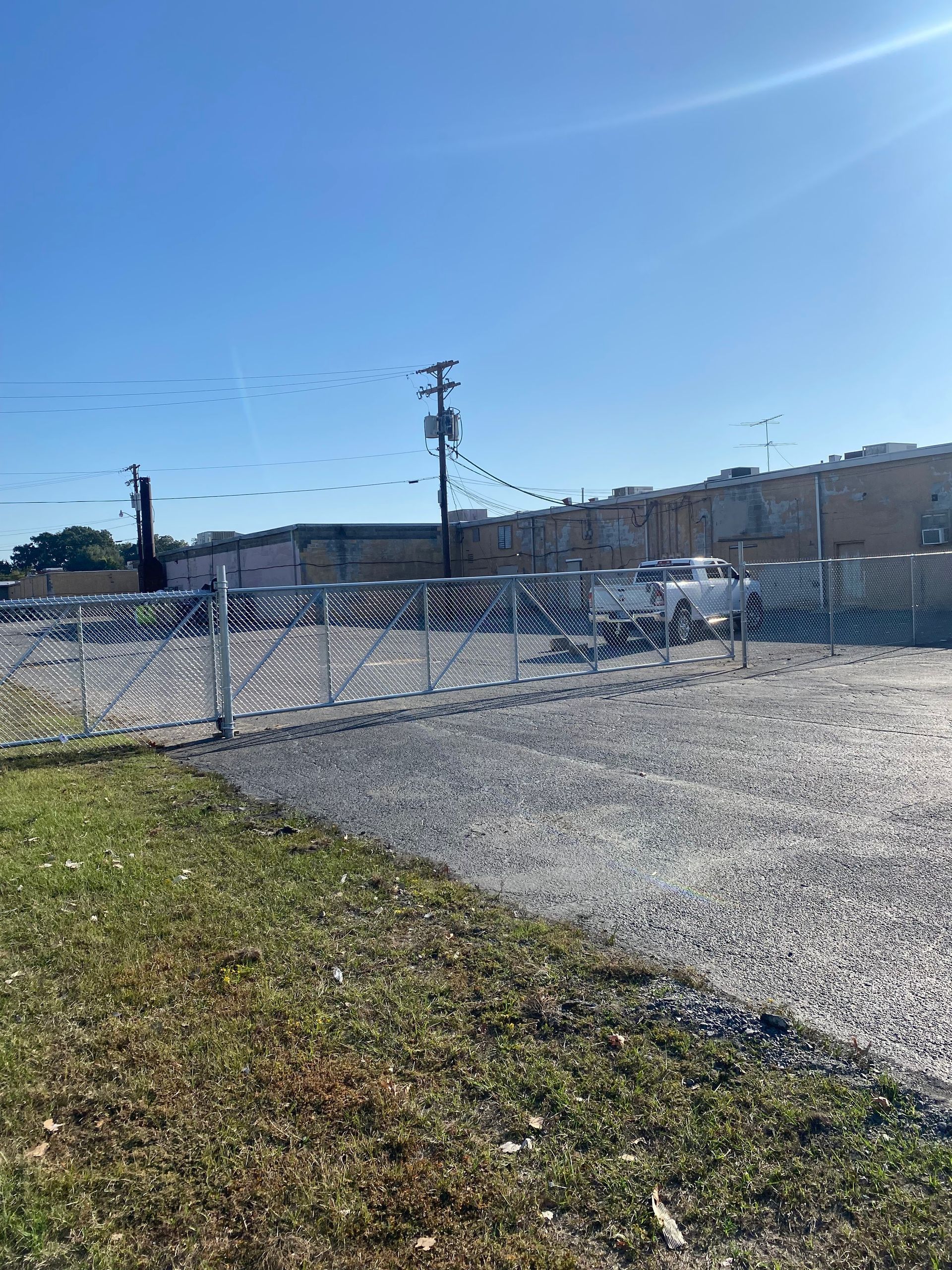 A chain-link fence encloses a gravel lot with a building in the background. Bright sky.
