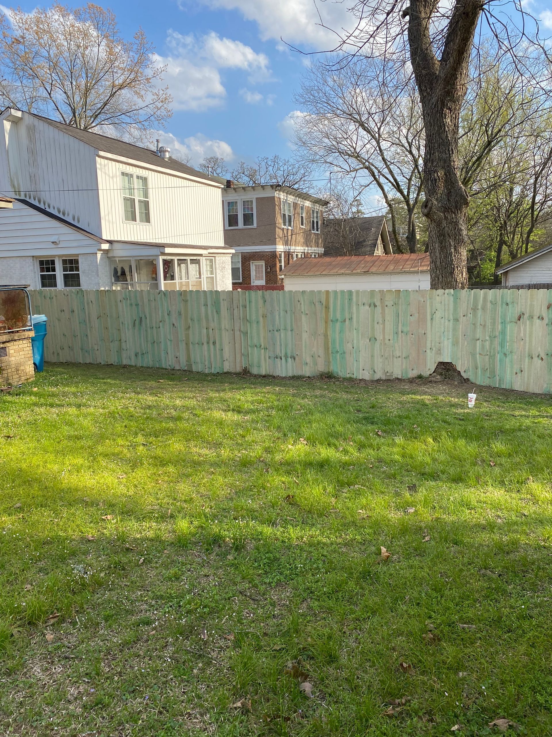 A newly built wooden fence in a grassy backyard, houses in background under a sunny sky.
