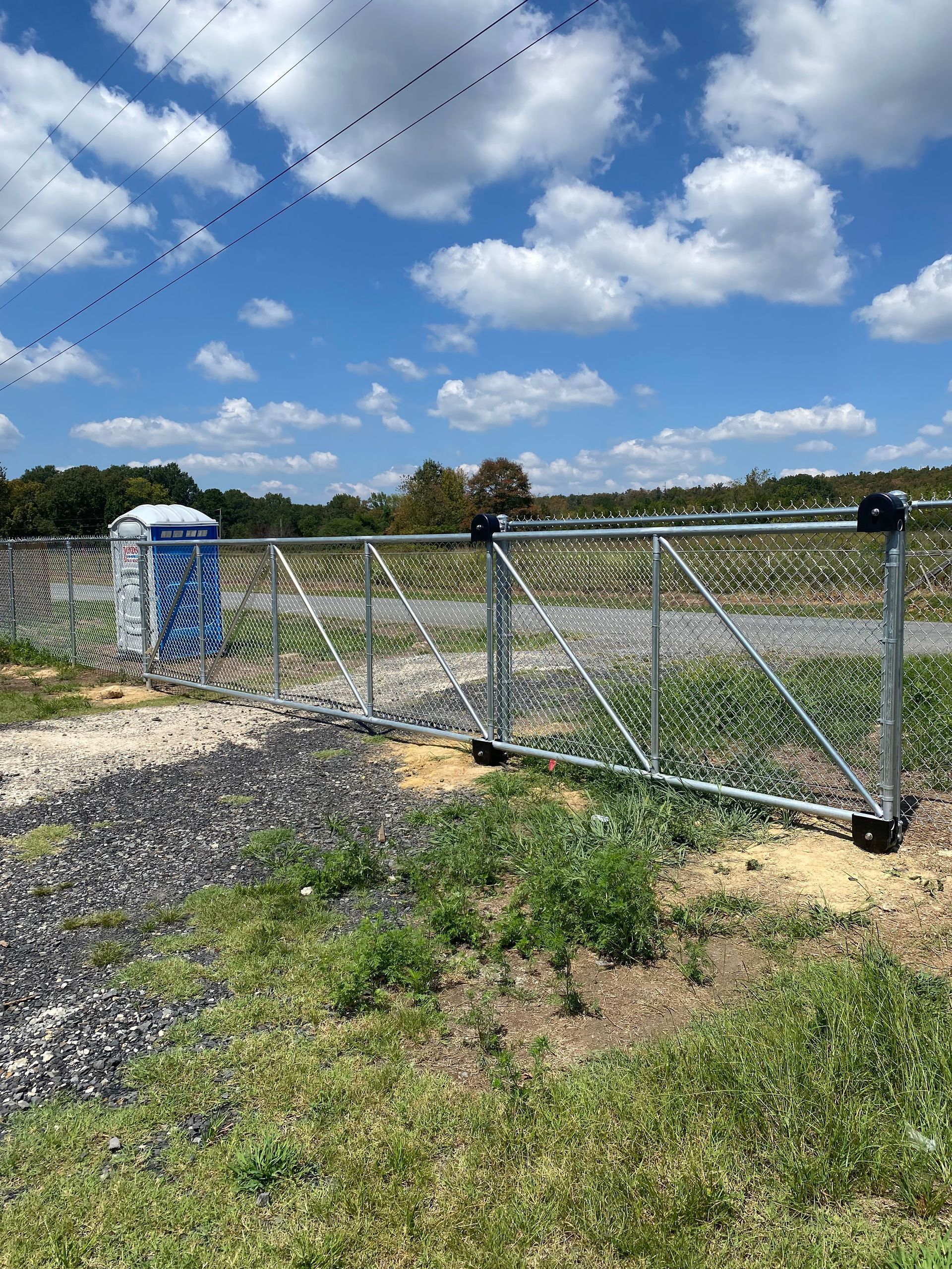 A portable toilet next to a metal gate on wheels in a grassy field, with a blue sky and clouds overhead.