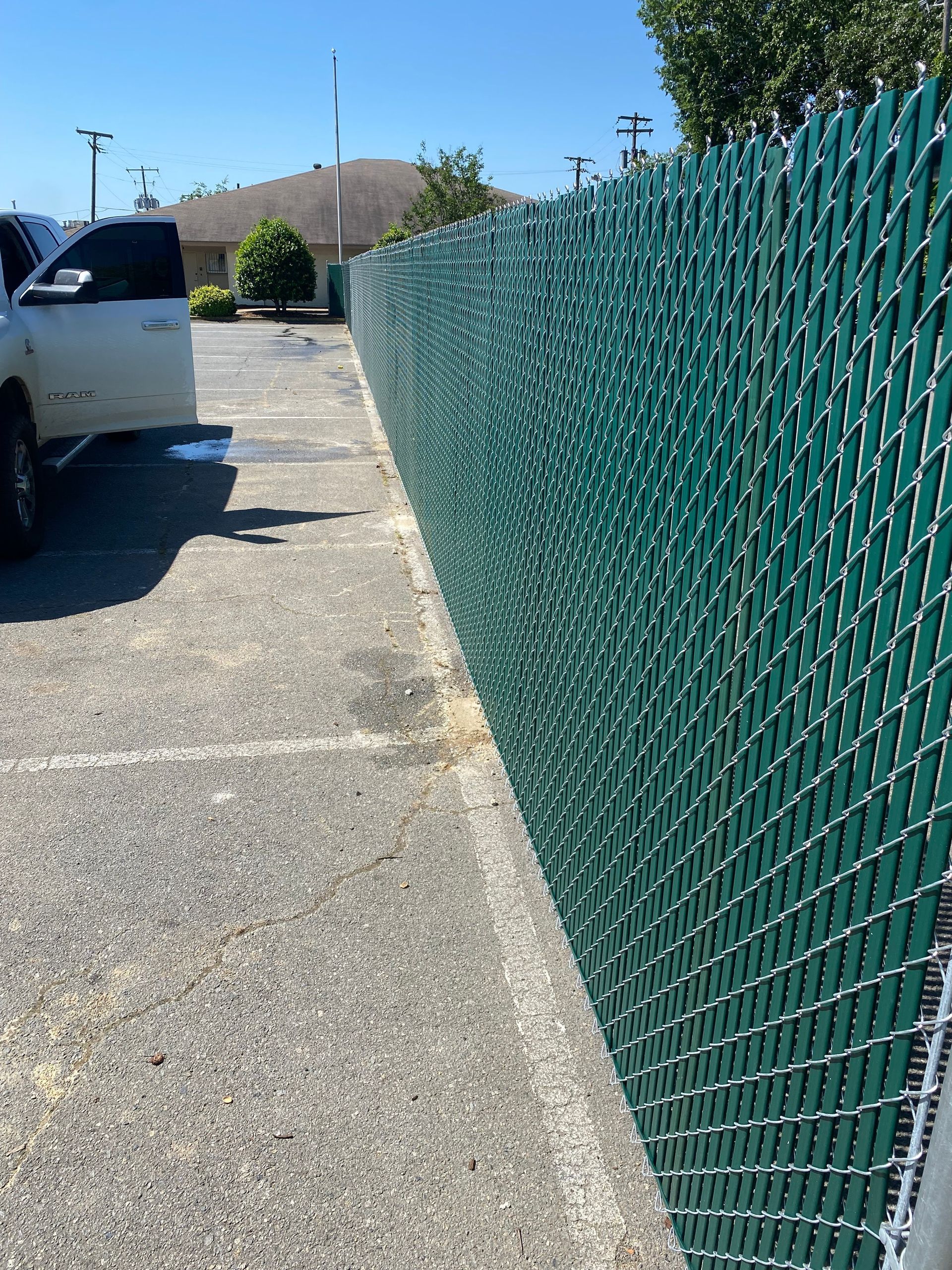 Green chain-link fence bordering a paved parking area on a sunny day. A white truck is partially visible.