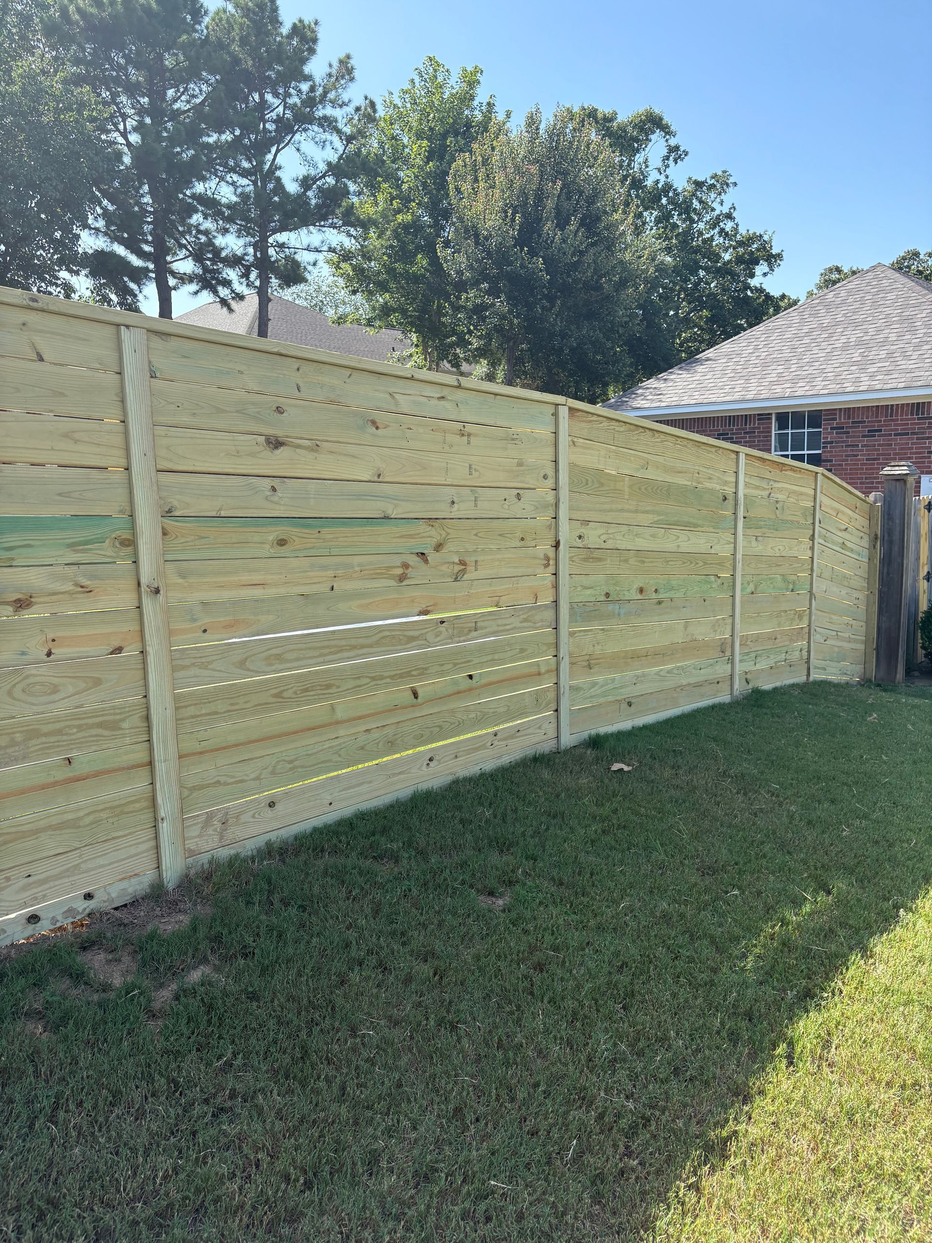 Wooden horizontal slat fence in a grassy yard, partially obscuring a house and trees under a blue sky.
