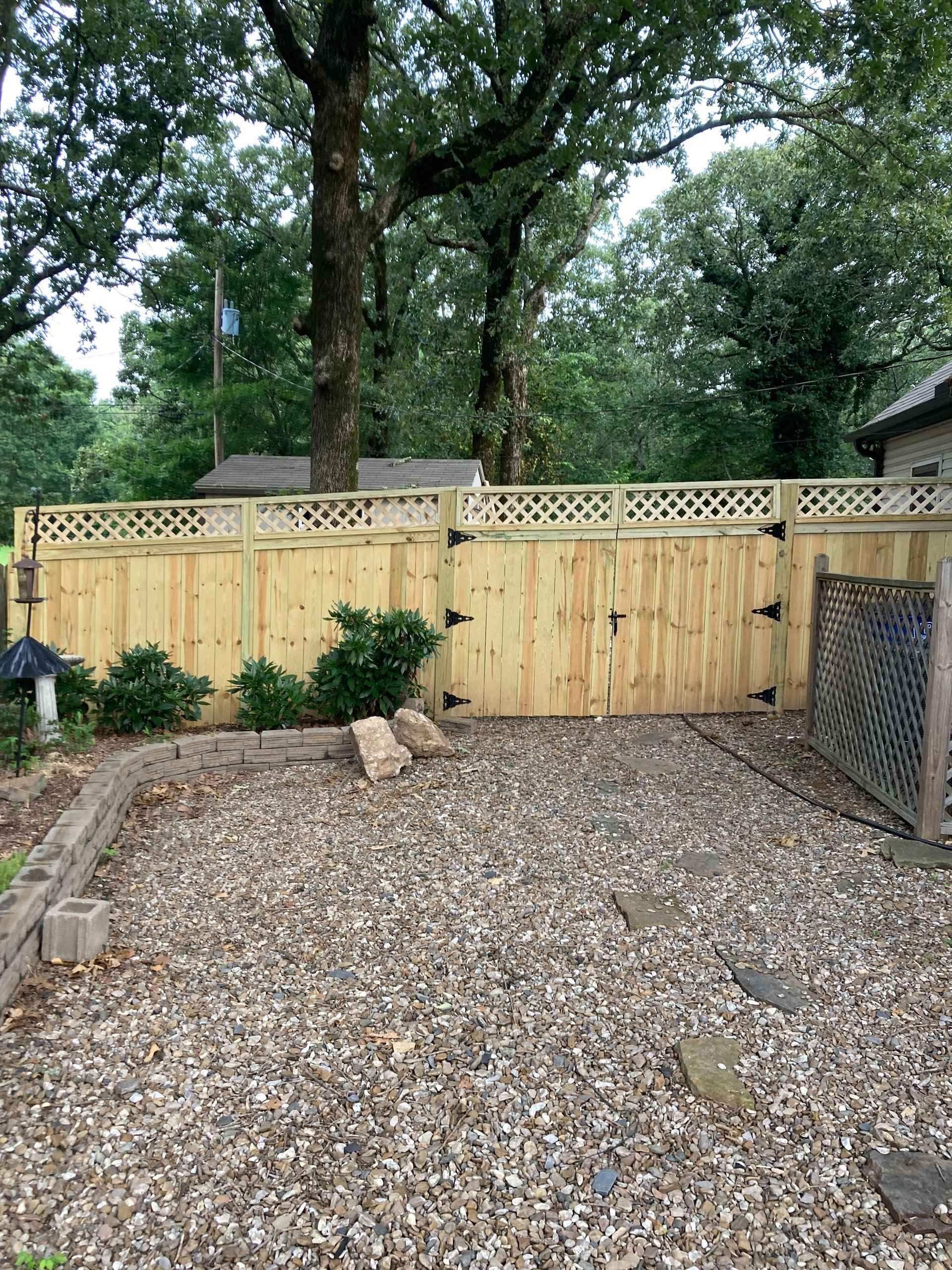 A wooden fence with a gate encloses a gravel yard. Trees and foliage are in the background.
