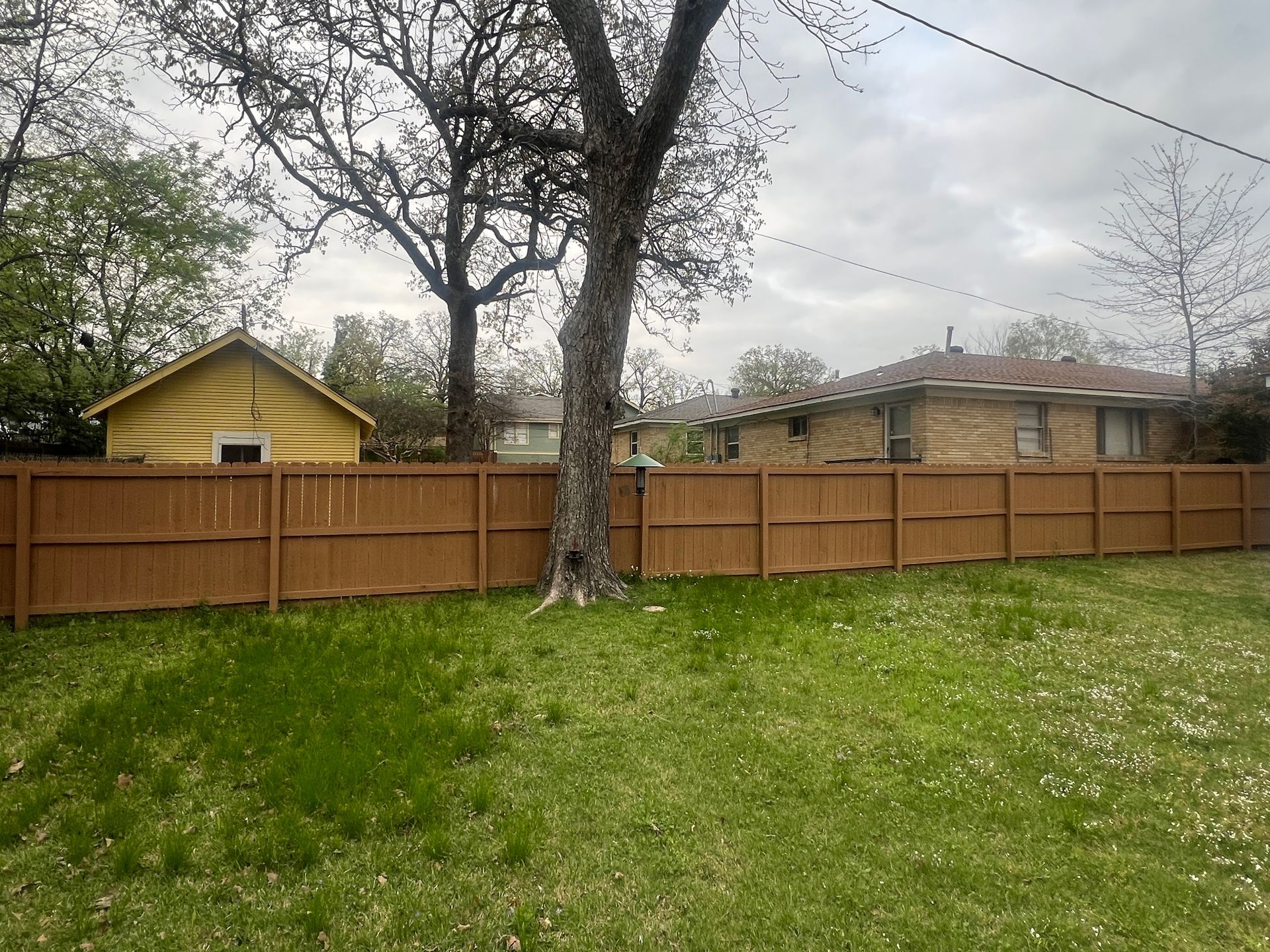 Brown wooden fence in a yard with green grass and a tree, houses in the background under a cloudy sky.
