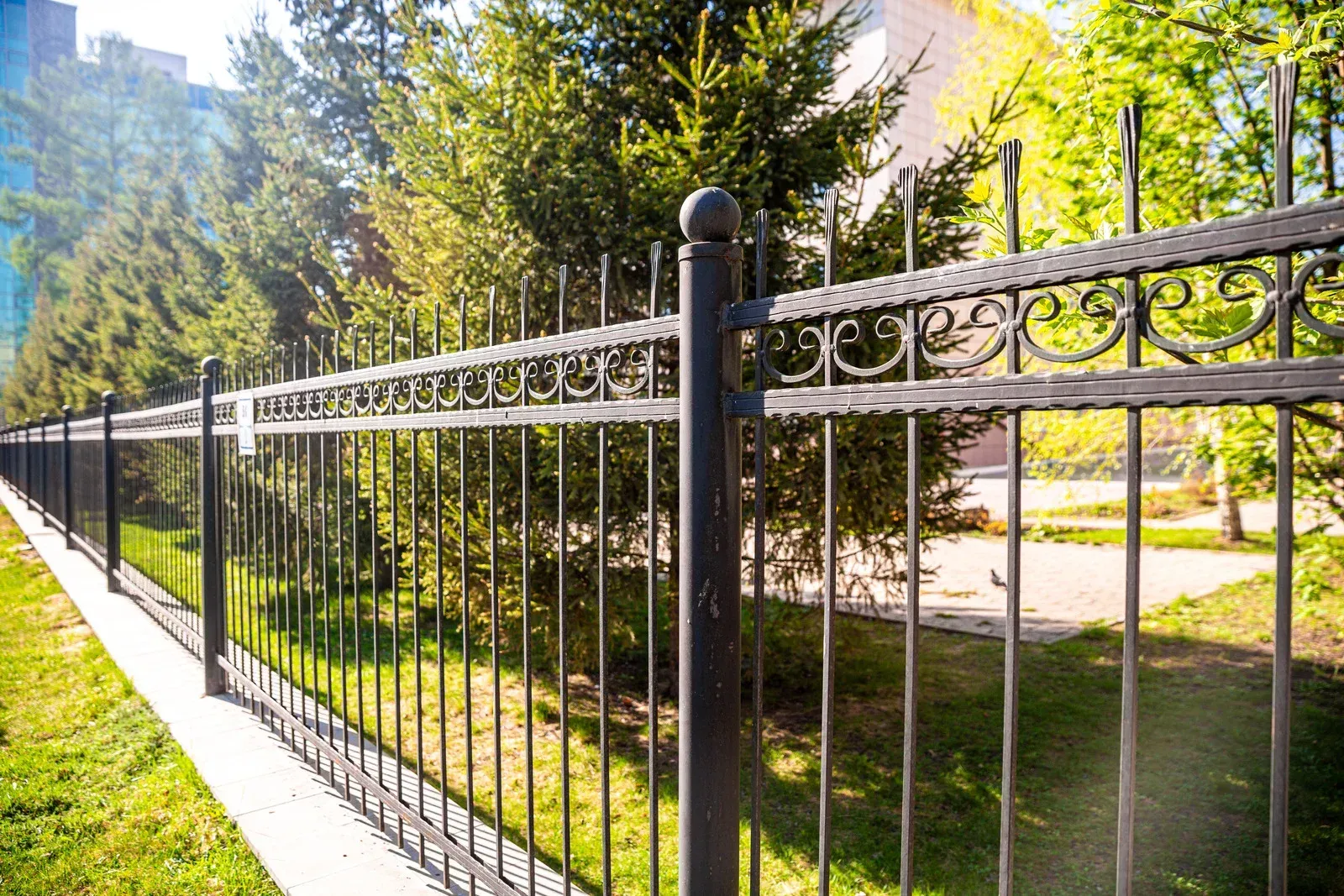 Black wrought iron fence with decorative scrollwork bordering a grassy area with trees.