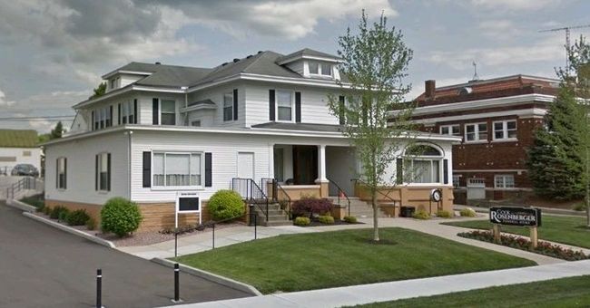 White two-story building with black shutters, brown trim, and a small front yard, beside a brick building.