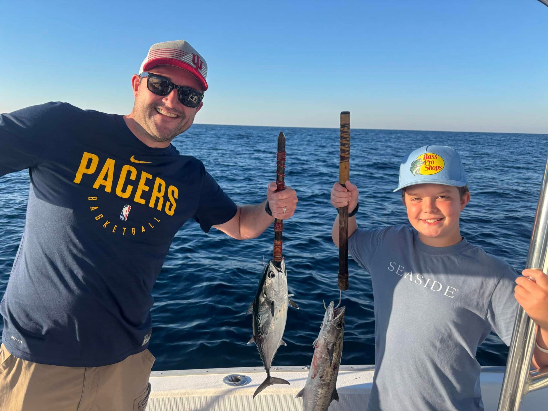 A smiling man and child on a boat hold up two fish they have caught in the deep blue ocean.