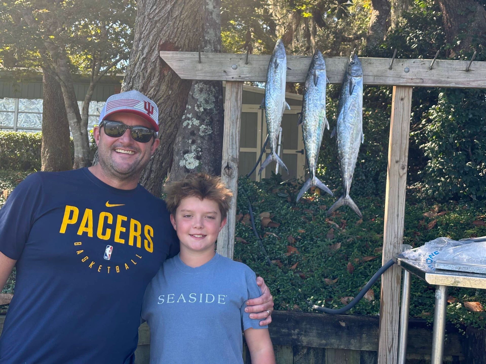 A smiling adult and child stand next to three fish hanging from a wooden rack in an outdoor, wooded setting.