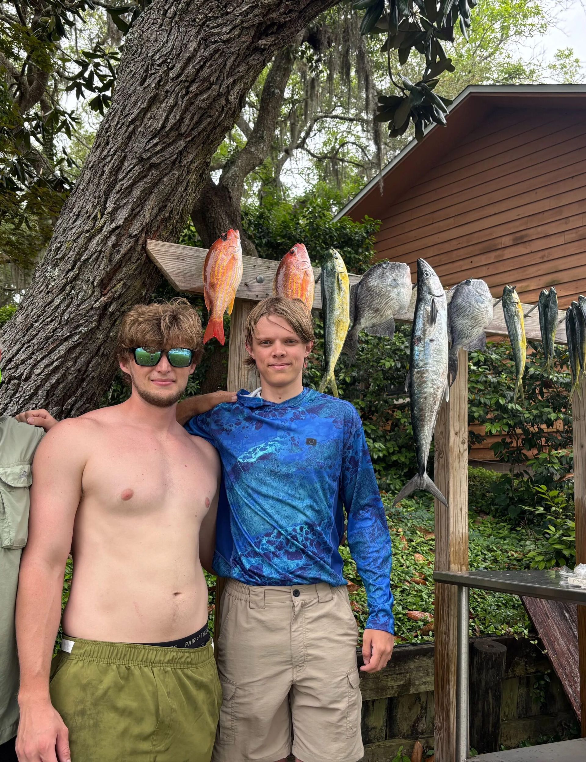 Two people standing in front of a rack of various caught fish hanging under a tree next to a wooden building.