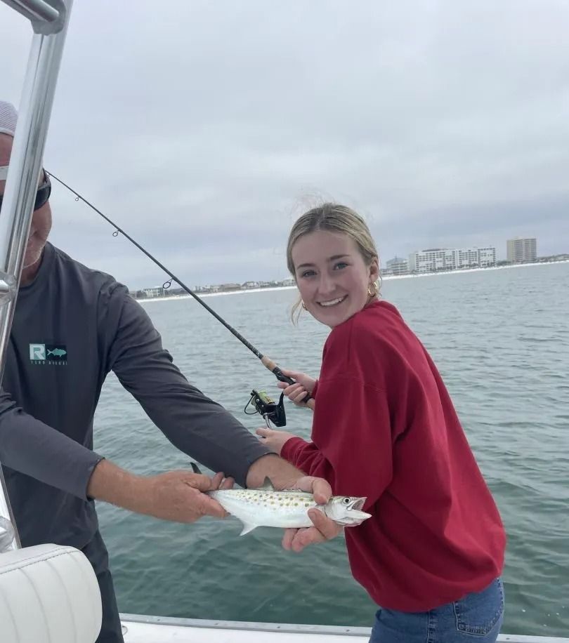 A person in a red sweatshirt and another person hold a spotted fish while fishing on a boat with the coast in view.