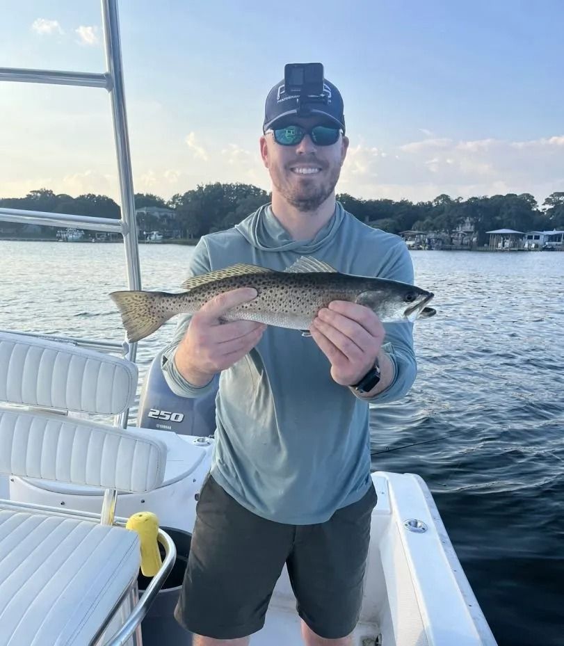 A person on a boat wearing a hat and sunglasses, holding a spotted seatrout in front of them with both hands.