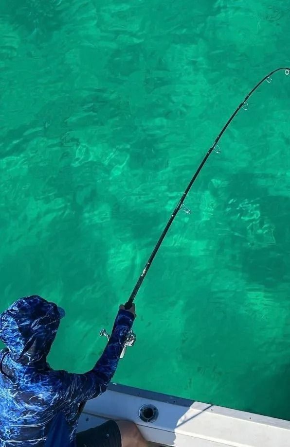 A person in a blue patterned shirt fishing from a boat over clear, bright turquoise water.