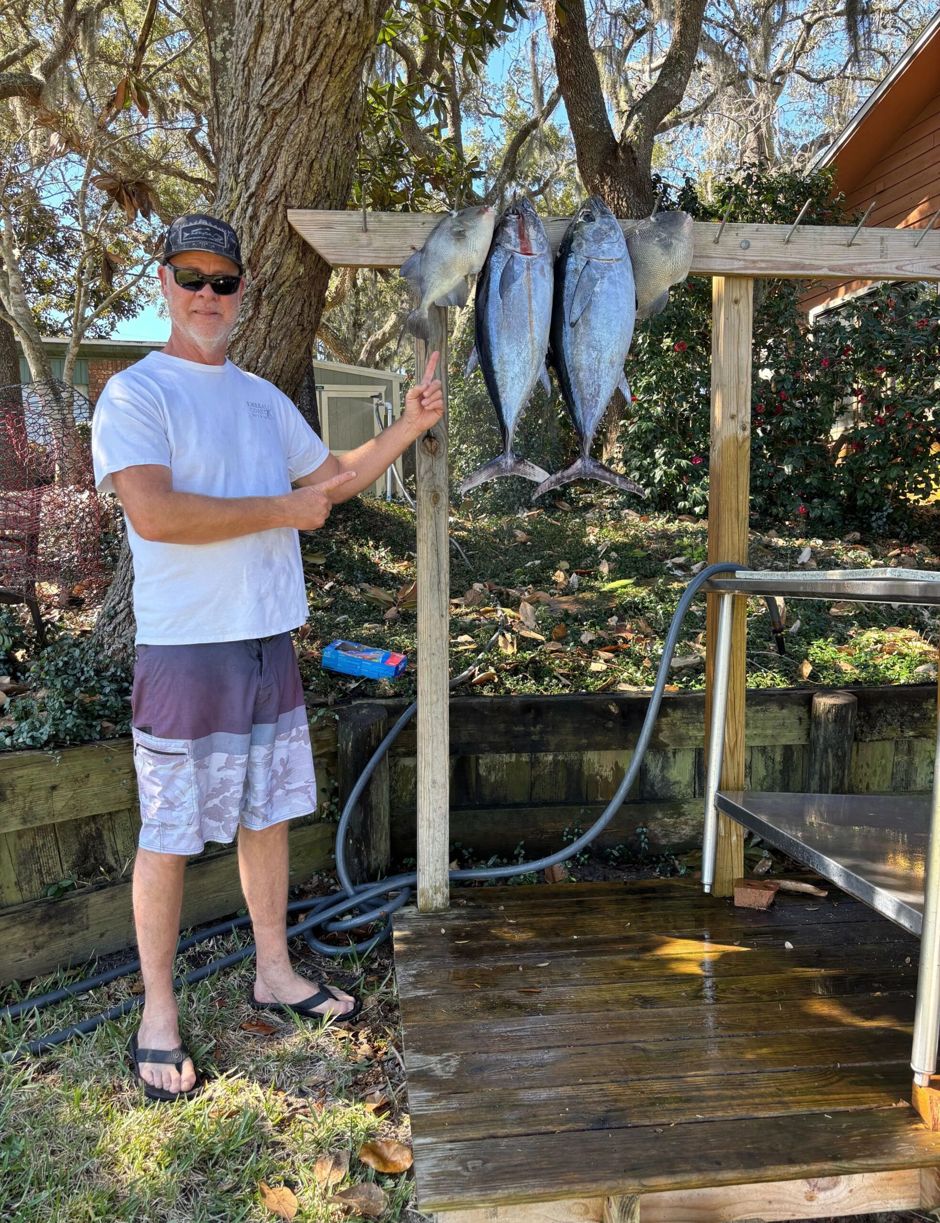 A man stands outdoors, gesturing toward four fish hanging from a wooden beam over a cleaning station.