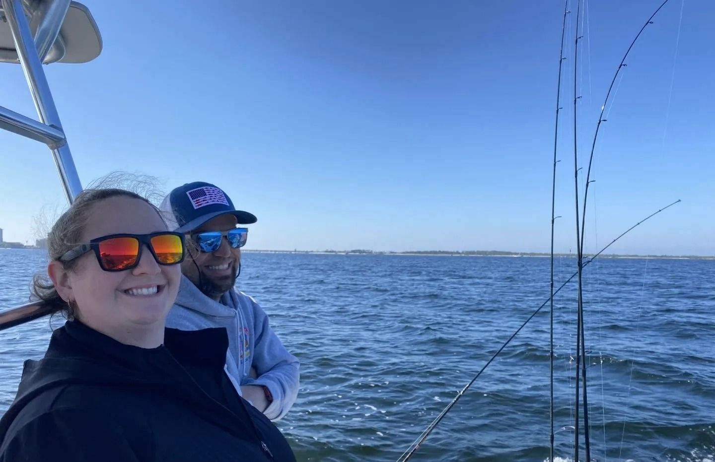 Two people wearing sunglasses and hats smile on a boat with fishing rods visible against a sunny ocean background.