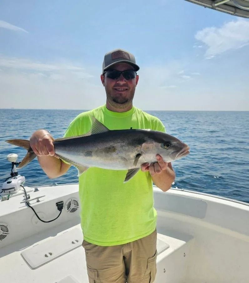 A man in a neon yellow shirt stands on a boat on the ocean, holding a fish he has caught.