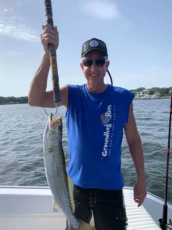 A person wearing a blue shirt, sunglasses, and cap holds a speckled trout fish hanging from a scale on a boat.