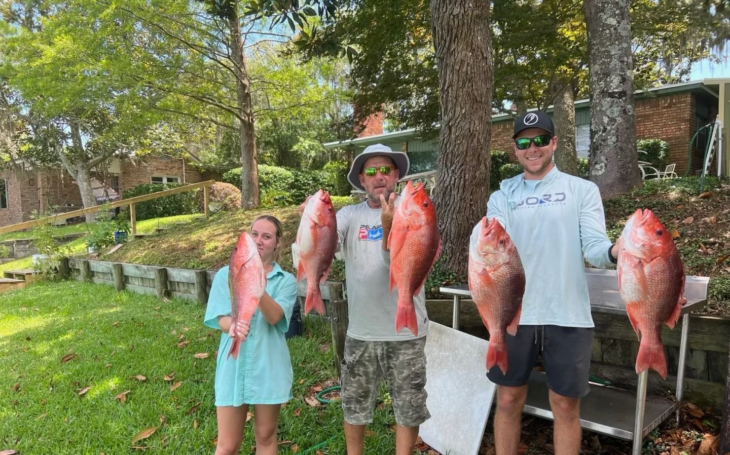 Three people stand outside in a grassy area, each holding a large red snapper fish, smiling toward the camera.