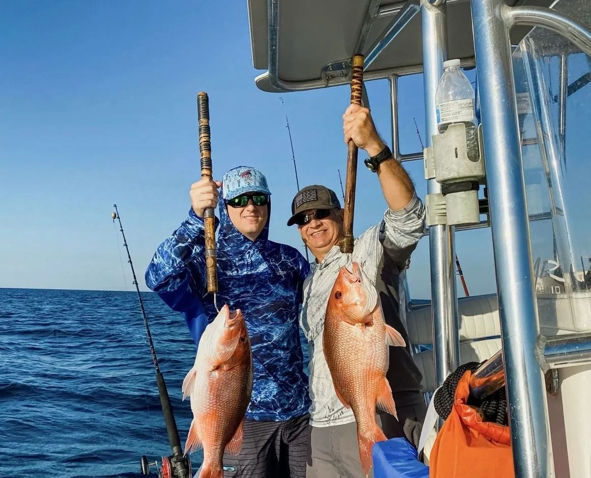Two people on a boat hold up large, caught red snapper fish against a bright blue sky and ocean background.