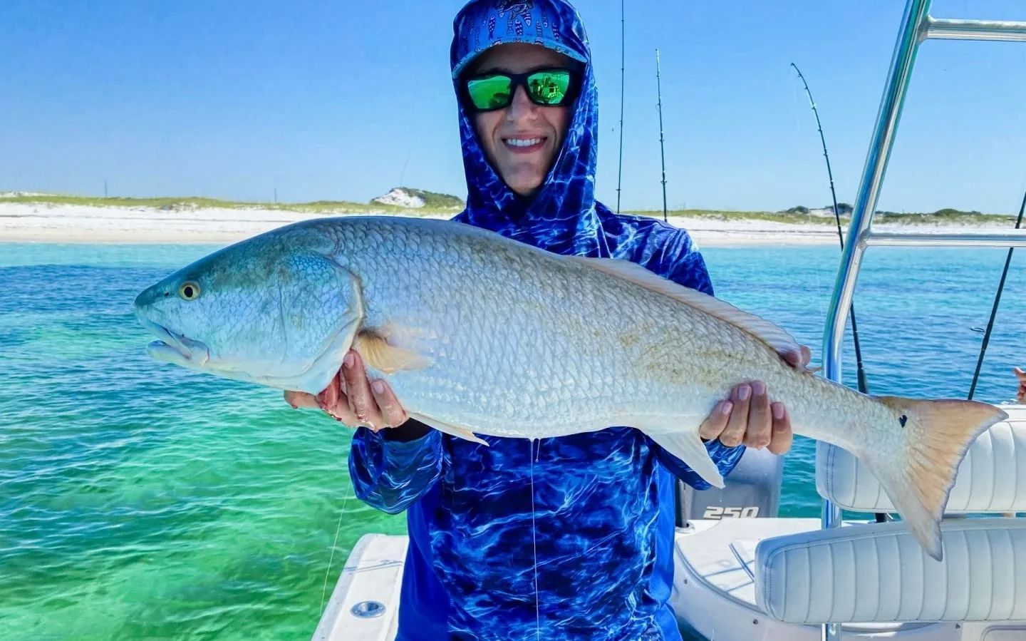 A person in a blue sun shirt and sunglasses holding a large, silvery fish on a boat in clear, shallow water.