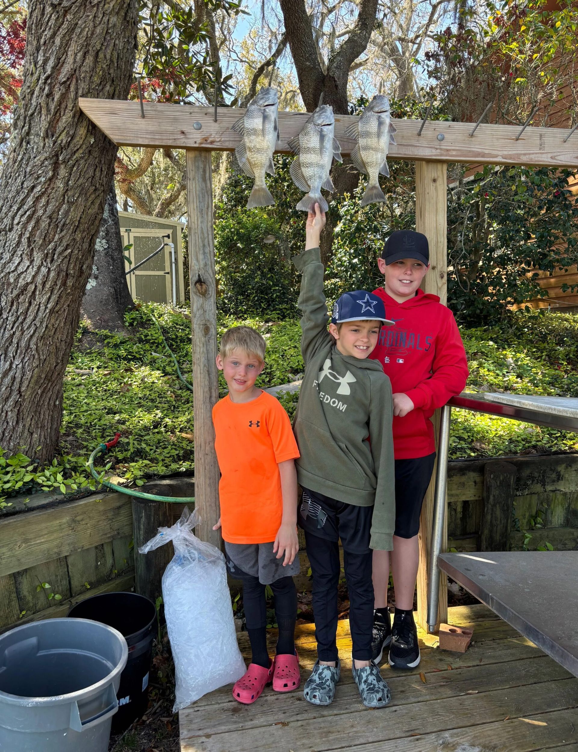 Three youths stand under a wooden rack holding three hanging fish in a wooded outdoor setting.