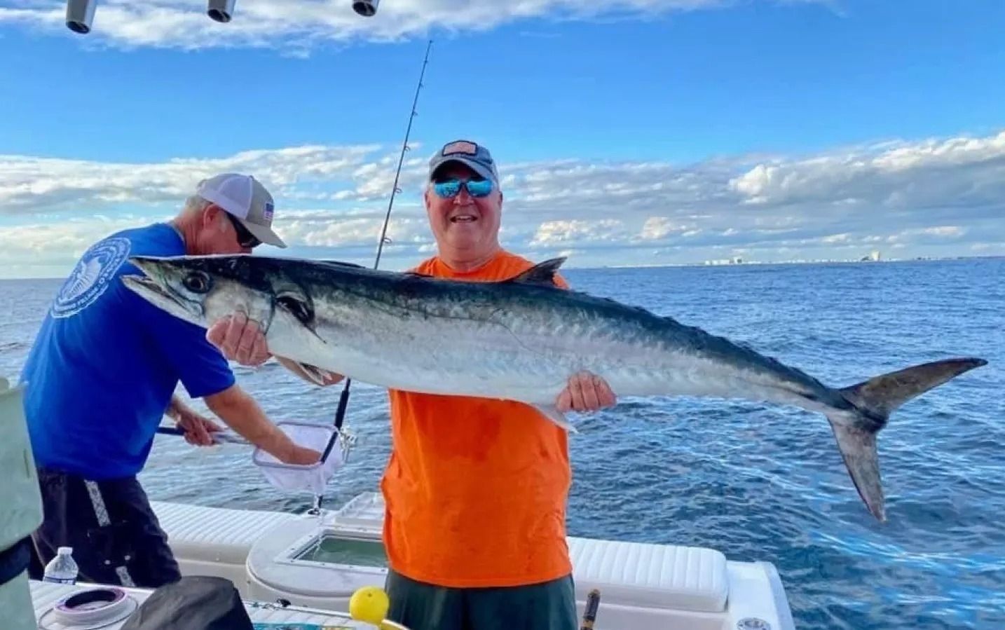 Two people on a boat, one in an orange shirt holding a large caught mackerel over the water under a clear sky.