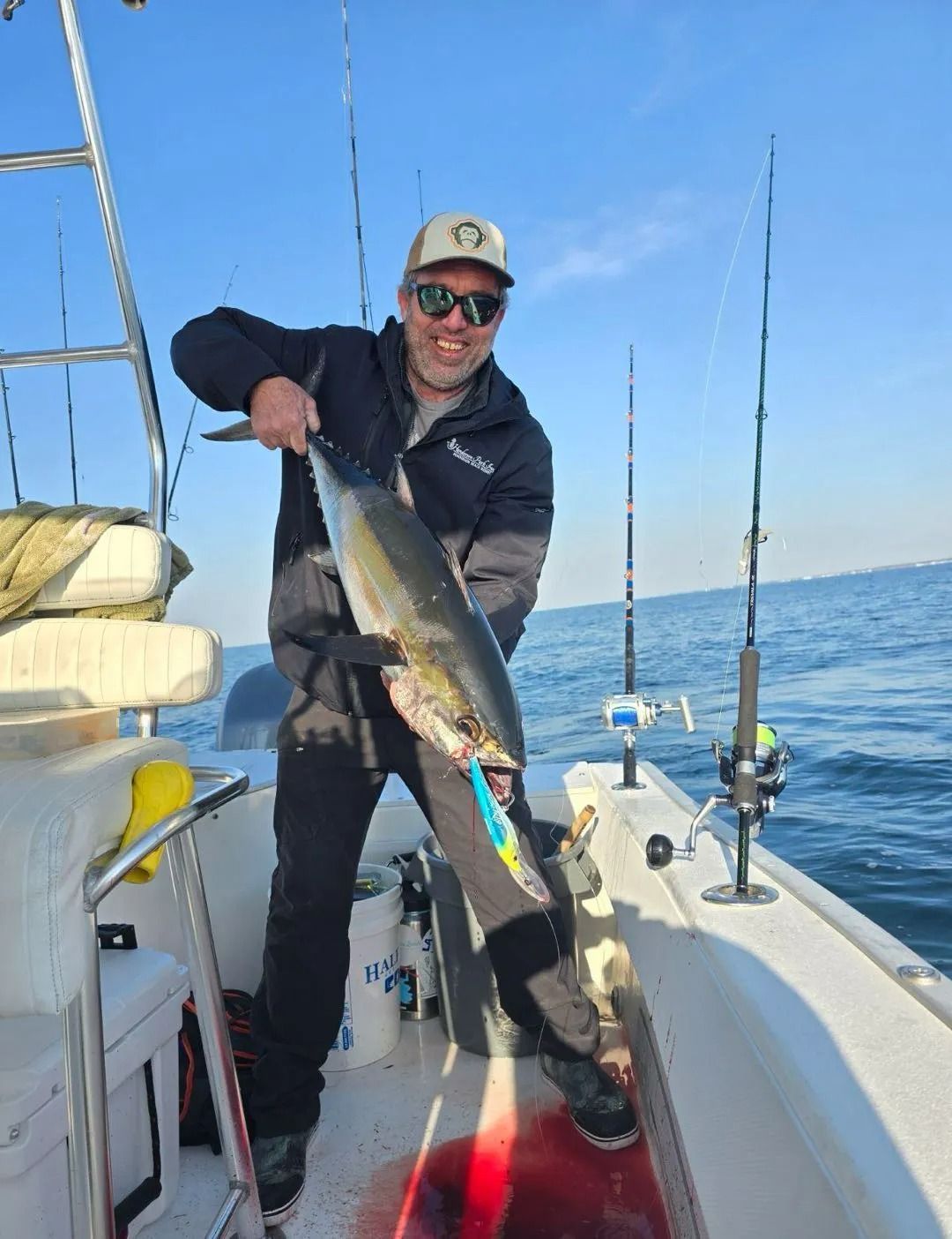 A smiling person on a boat holds up a freshly caught tuna with a blue lure in its mouth against a clear blue sky.