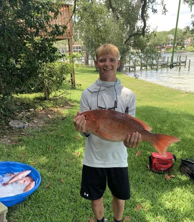 A smiling person holds a large, red snapper fish outdoors by a grassy waterfront, with more fish in a blue bin nearby.