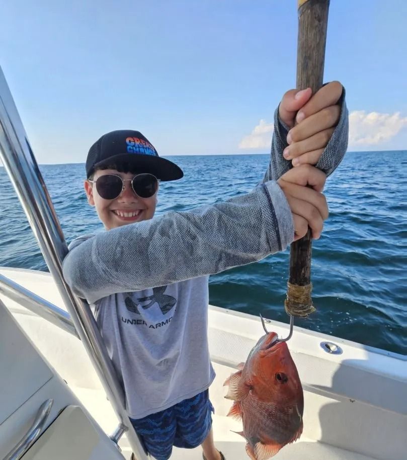 A person on a boat smiles while holding up a freshly caught red snapper on a gaff hook against an ocean background.