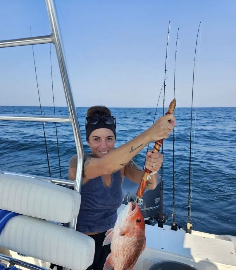 A woman on a boat smiles while holding a large red snapper fish on a metal hook, with fishing rods in the background.