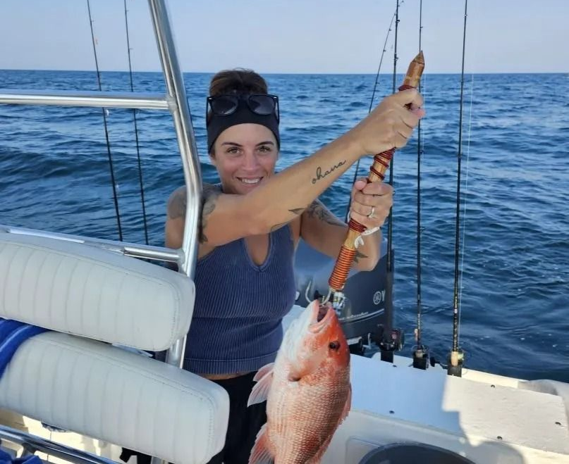 A person smiling on a boat, holding a fishing gaff with a red snapper caught on the hook against a blue ocean background.