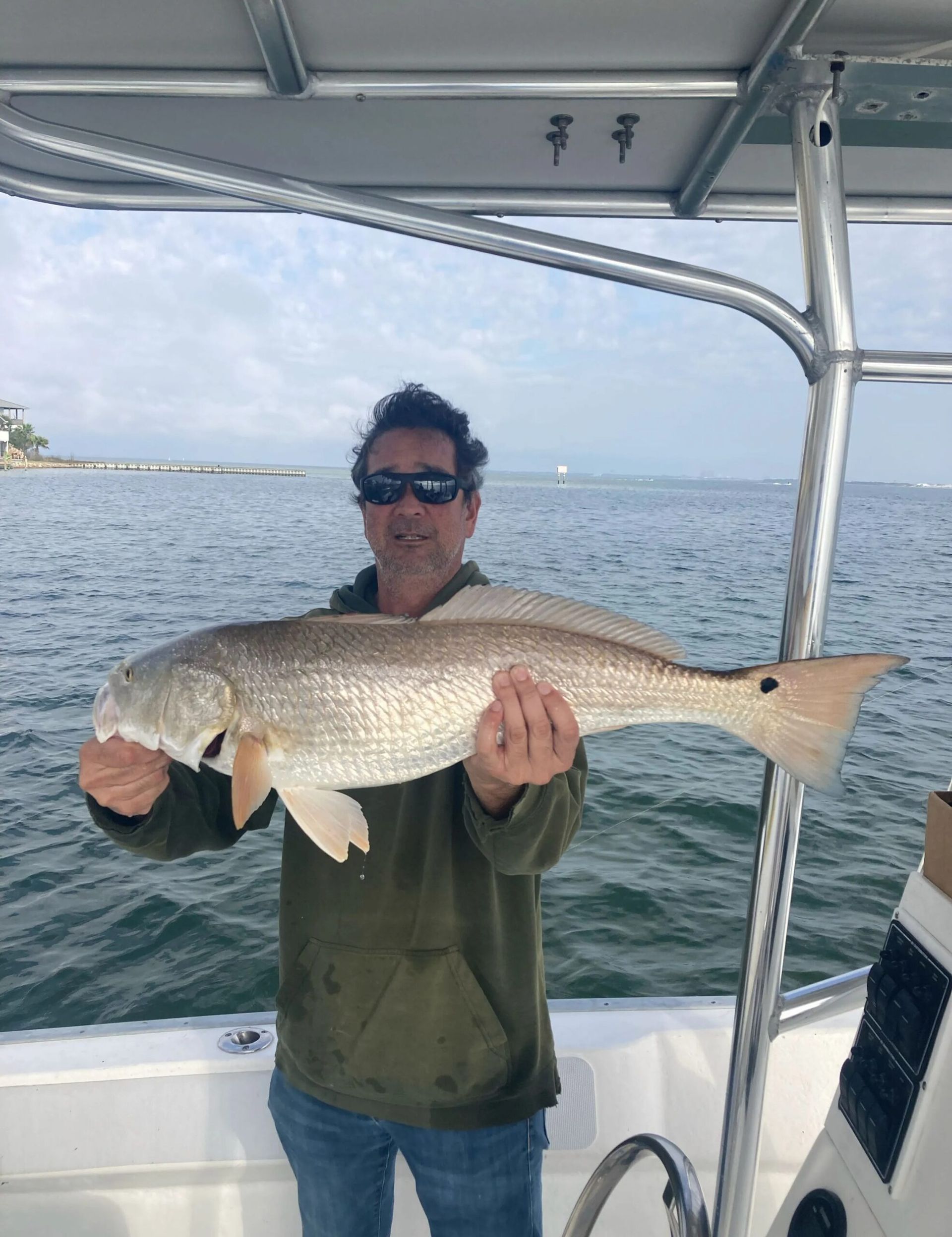 A person wearing sunglasses and a green hoodie holds a large fish while standing on a boat on the water.