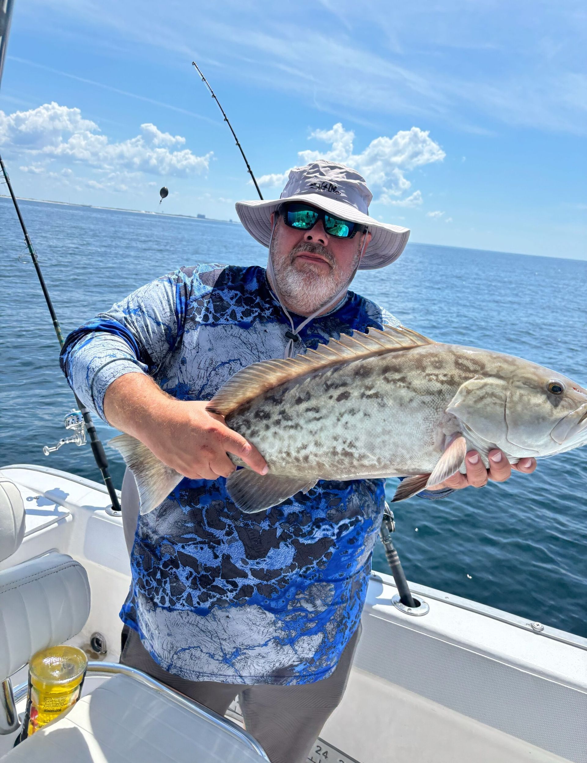 A person in a blue patterned shirt and hat holds a large, spotted fish on a boat on the open ocean.