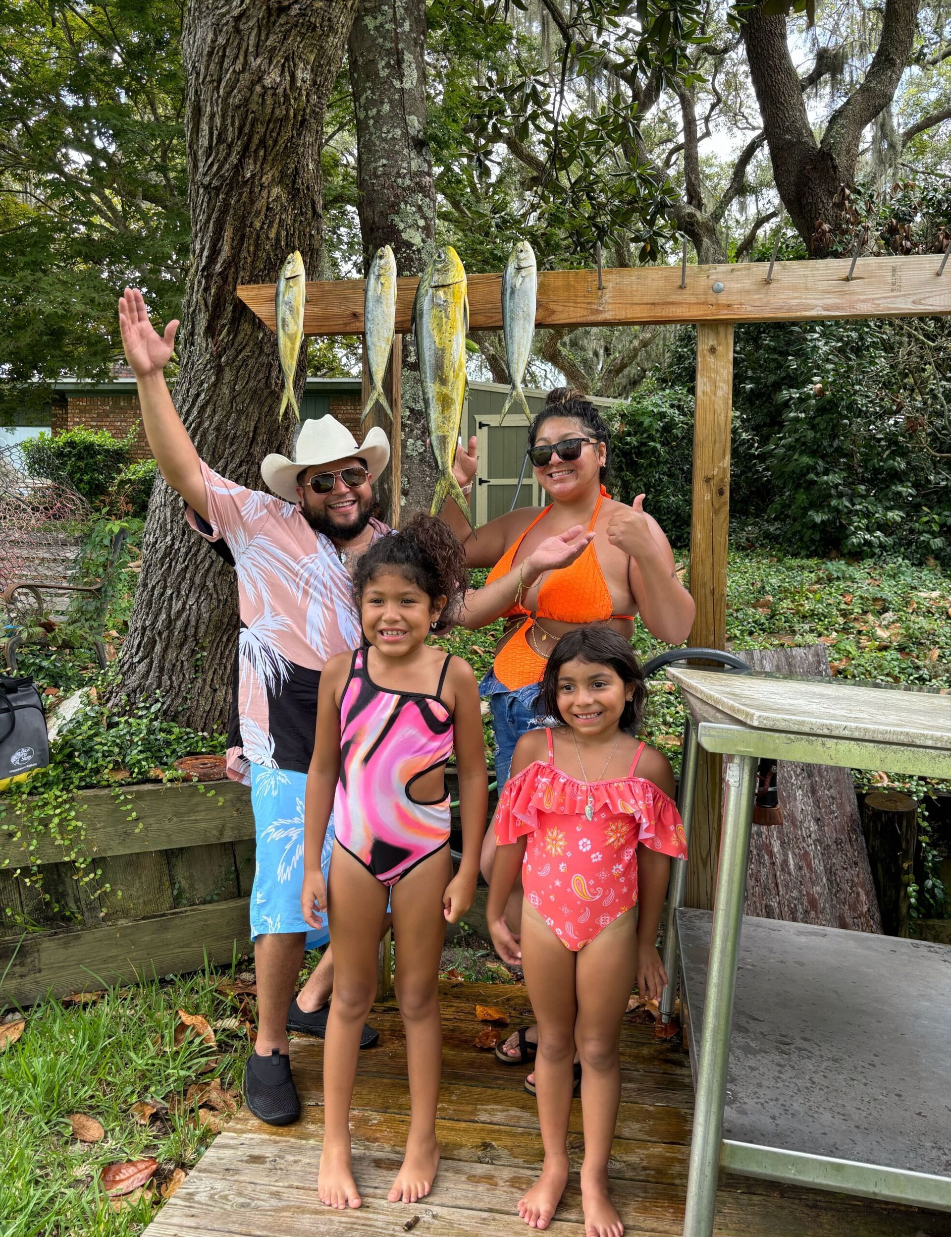 A family stands outside, smiling and posing by a wooden rack hanging with four fish, near a metal table in a yard.