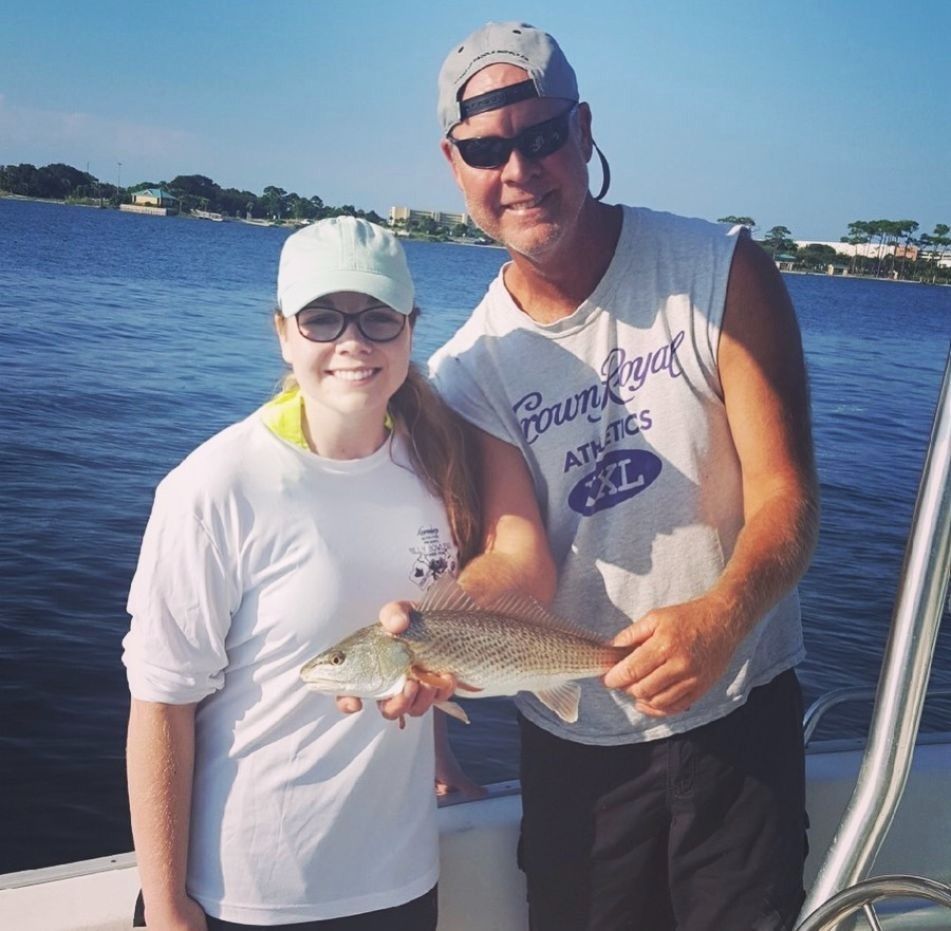 A smiling person and an adult holding a freshly caught fish on a boat on the water under a clear blue sky.