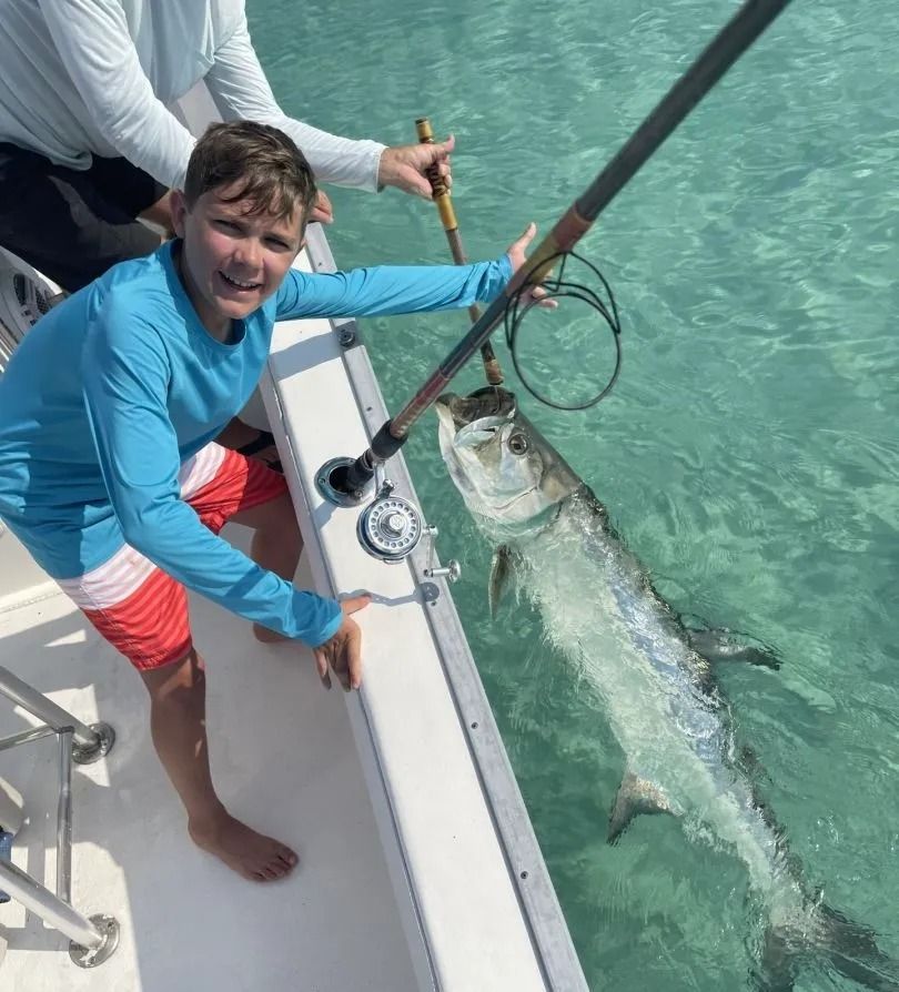 A smiling person in a blue shirt holds a fishing rod over the side of a boat, reeling in a large, silver tarpon fish.