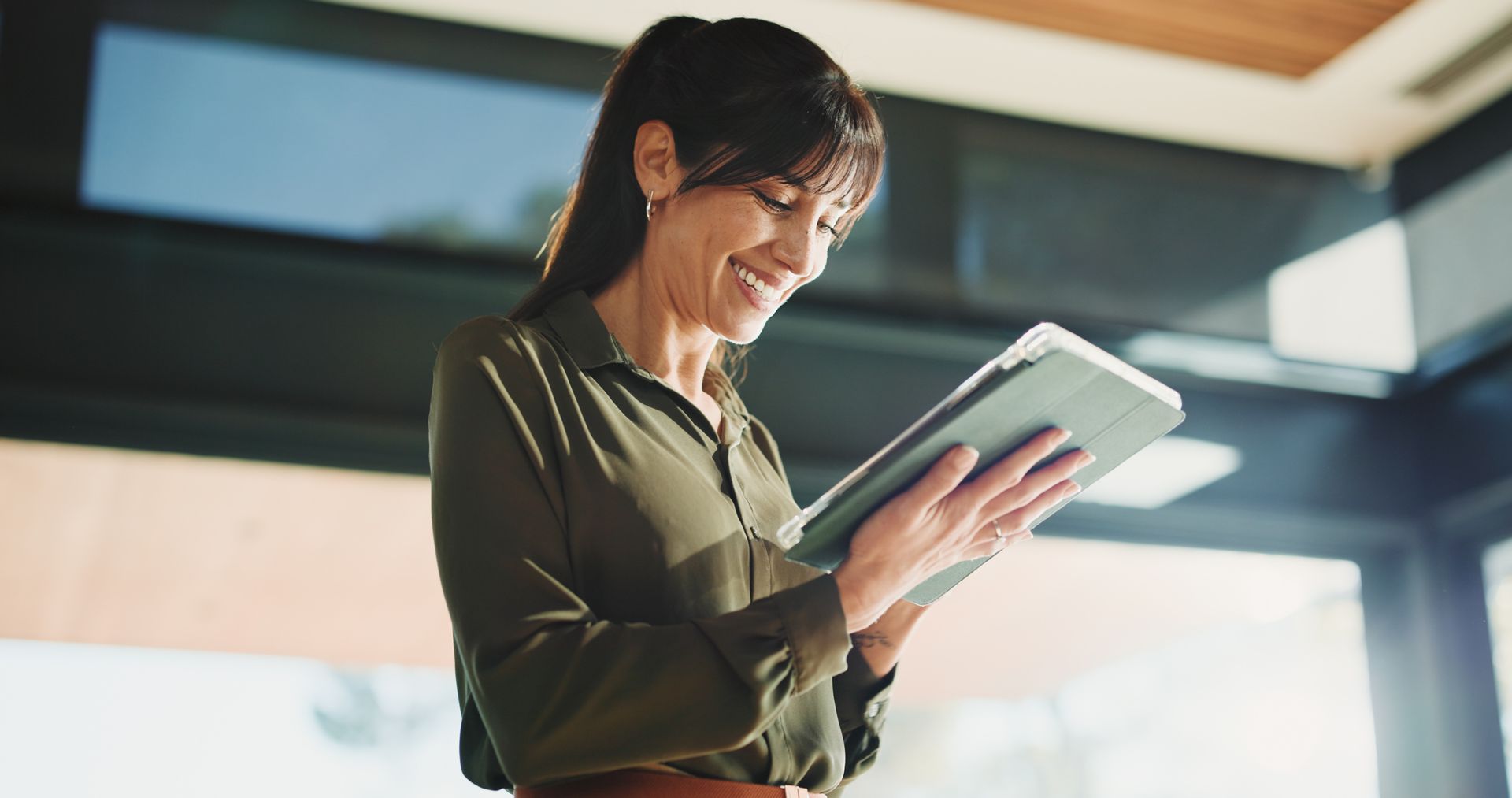 A smiling professional in an olive green shirt stands in an office setting, holding and looking at a digital tablet.