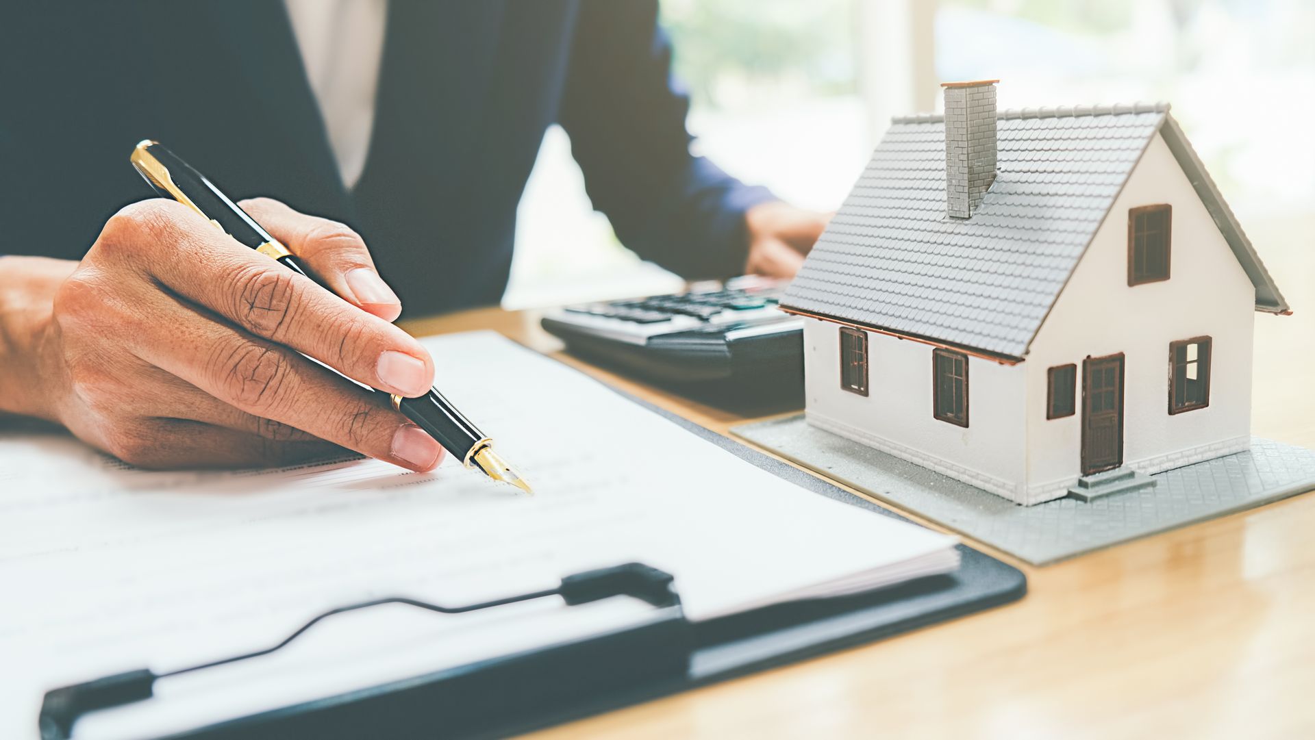 A person in a business suit writes on a document next to a small house model and a calculator on a wooden desk. A person in a business suit writes on a document next to a small house model and a calculator on a wooden desk.