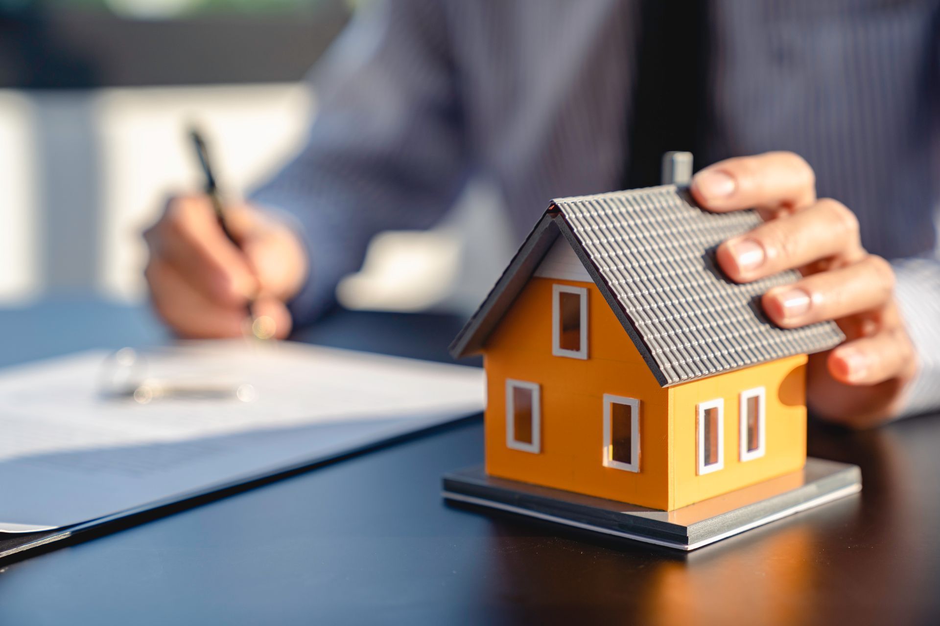 A person’s hand rests on a small orange model house on a desk while they write on a document nearby.