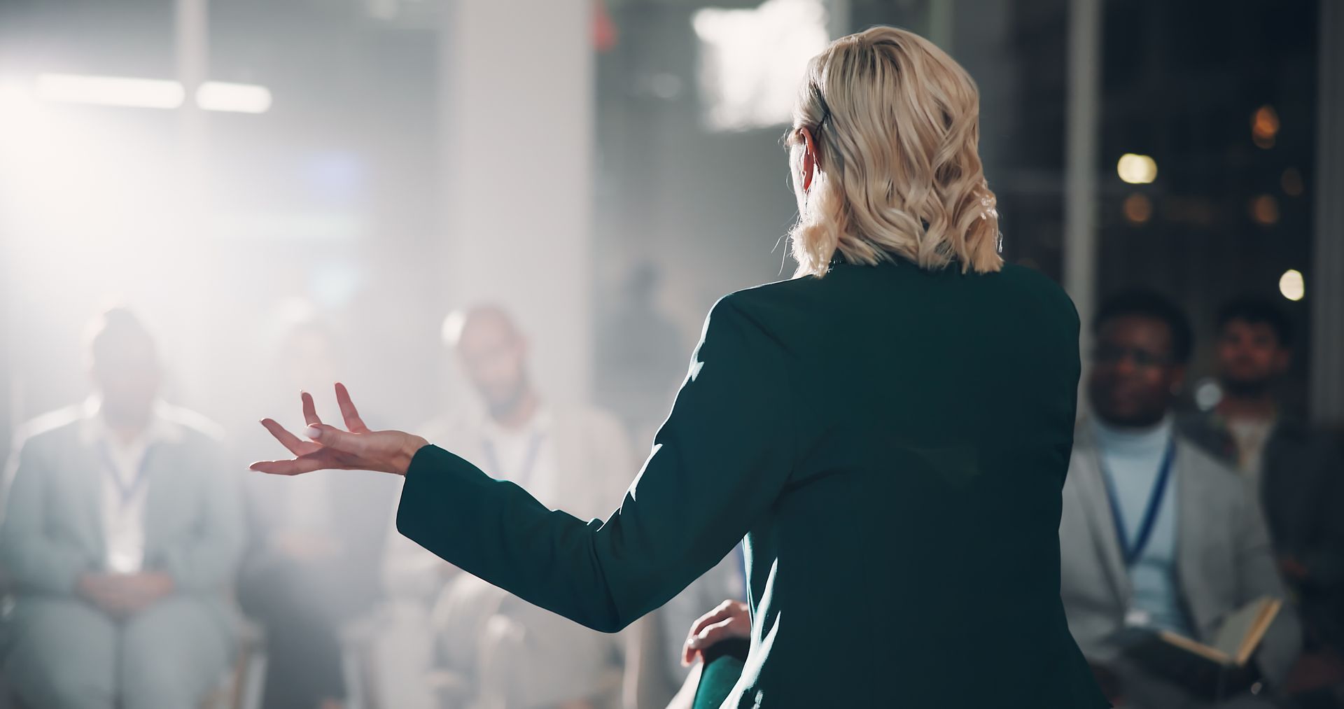 A person in a dark suit stands with their back to the camera, gesturing while addressing a blurred audience in a room.