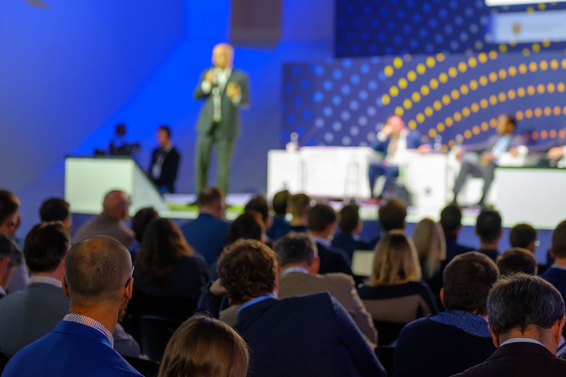 A speaker stands on a stage before an audience at an indoor conference with blue lighting and geometric background panels.