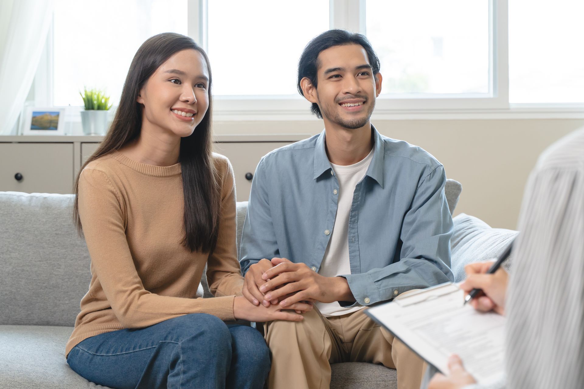 Two people sit on a couch, holding hands, talking with a therapist in a bright living room.