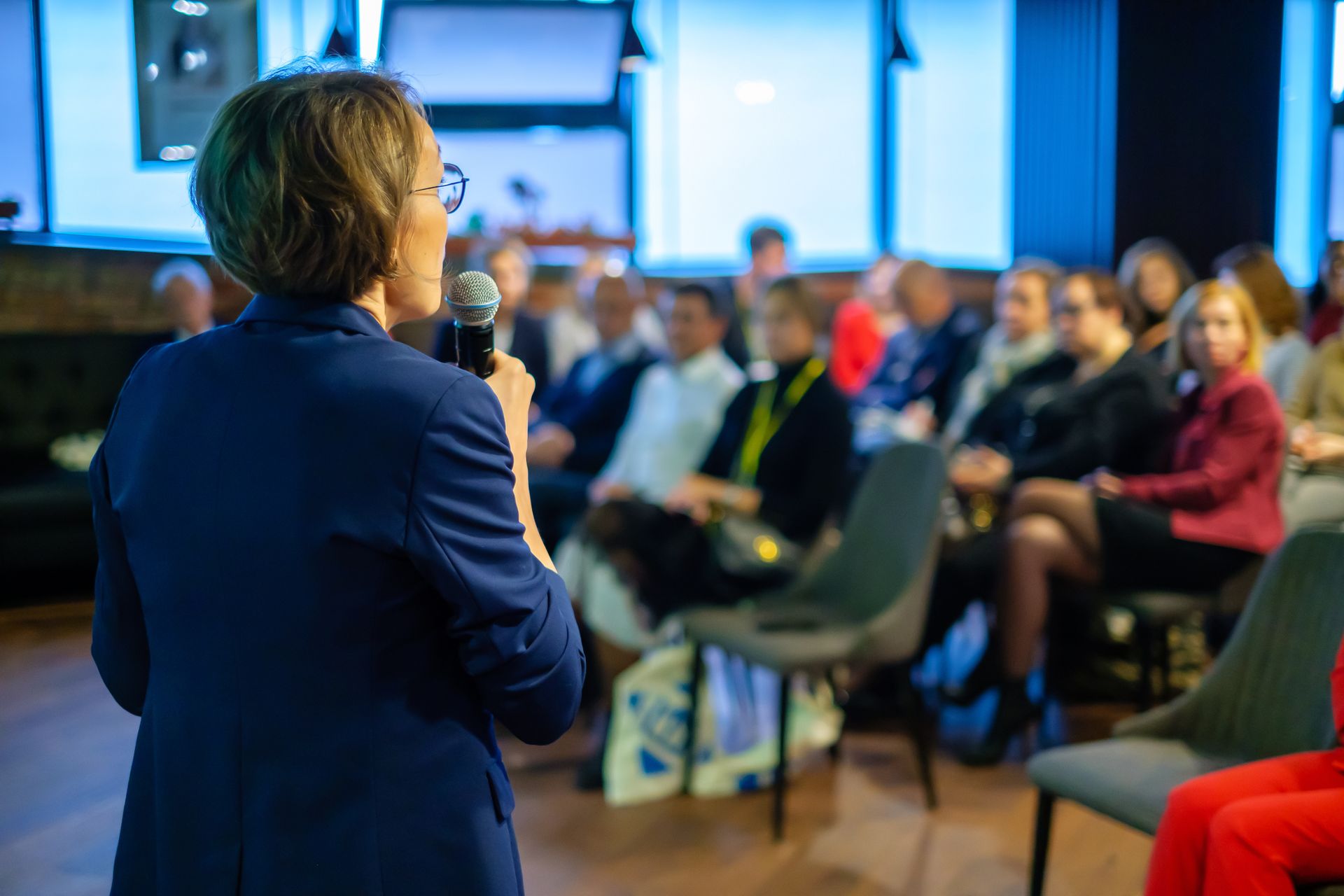 A speaker in a blue blazer stands before an audience in a room with blue-lit windows, holding a microphone.